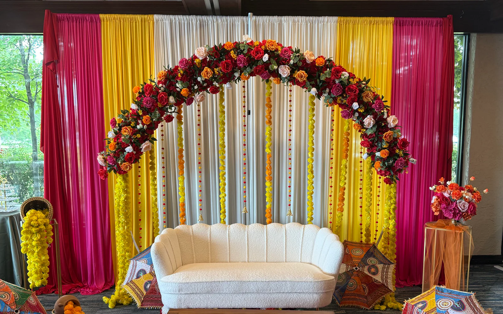 Colorful South Asian celebration decor with floral arch, pink and yellow draping, white sofa, and decorative umbrellas.